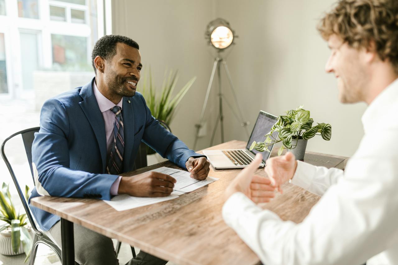 two people sitting at a desk smiling and explaining something to one another