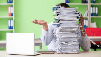 A person at an office desk is partially obscured by a large stack of paperwork, holding a phone beside an open laptop, with green walls and shelves of binders behind them.