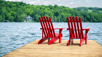 Two red Muskoka chairs sit on a wooden dock overlooking a calm lake, with trees and hills in the background and kayaks tied at the edge.