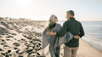 Two people walk side by side along a sandy beach near the shoreline, wearing jackets with their arms around each other, with the ocean and a clear sky in the background.