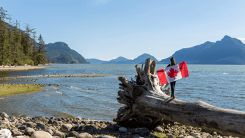 Person standing on a large driftwood log by a lakeshore, holding a Canadian flag with mountains and forest in the background.