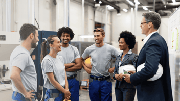 Six people standing together in an industrial workspace, wearing work and safety clothing, gathered around a person holding a clipboard and hard hat, with machinery in the background.