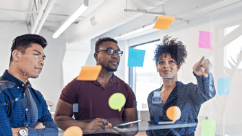 Coworkers collaborate in an office using a glass board with colorful sticky notes.