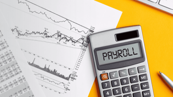 A calculator displaying the word “PAYROLL” sits on a yellow surface beside printed financial charts and graphs showing lines and data points.
