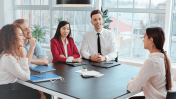 Five people sit around a conference table in a bright office space, with laptops and notebooks on the table, engaged in a discussion near large windows and indoor plants.