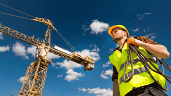A construction worker wearing a high-visibility safety vest and hard hat stands holding a coiled rope, with a tower crane and blue sky with clouds in the background.