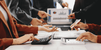 many hands working at a long table, using a calculator, writing notes and pointing to a screen with a report on it