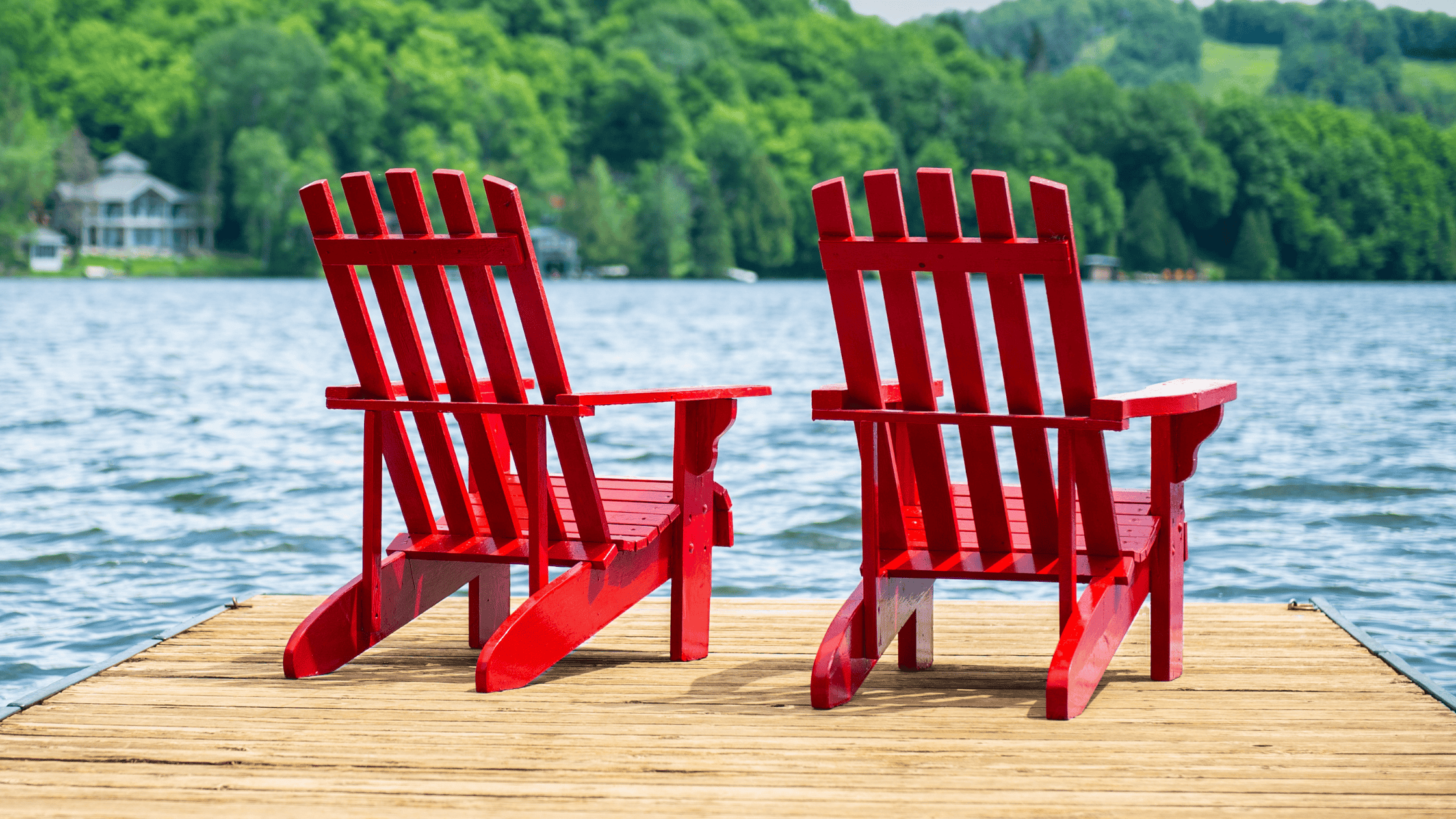 Two red Muskoka chairs sit on a wooden dock overlooking a calm lake, with trees and hills in the background and kayaks tied at the edge.