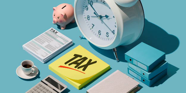 Tax‑themed workspace with a piggy bank, clock, coffee, tax documents, calculator, sticky notes labeled “TAX,” and blue binders on a light blue background.