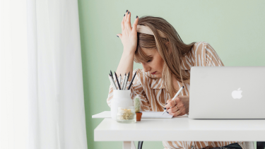 Person working at a desk with a laptop and paperwork, resting a hand on their head while reviewing notes, illustrating the challenges of managing tax or administrative tasks.