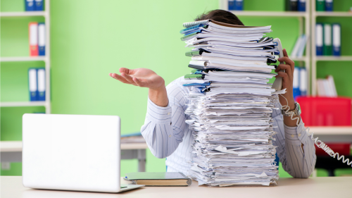 A person at an office desk is partially obscured by a large stack of paperwork, holding a phone beside an open laptop, with green walls and shelves of binders behind them.