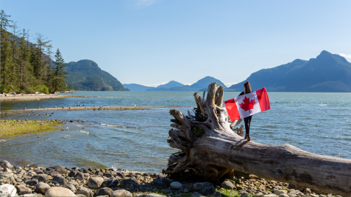 Generating response&nbsp; Copilot said:  A Canadian flag is displayed on a large driftwood log along a rocky shoreline, with mountains and a coastal inlet stretching into the distance under a clear blue sky.