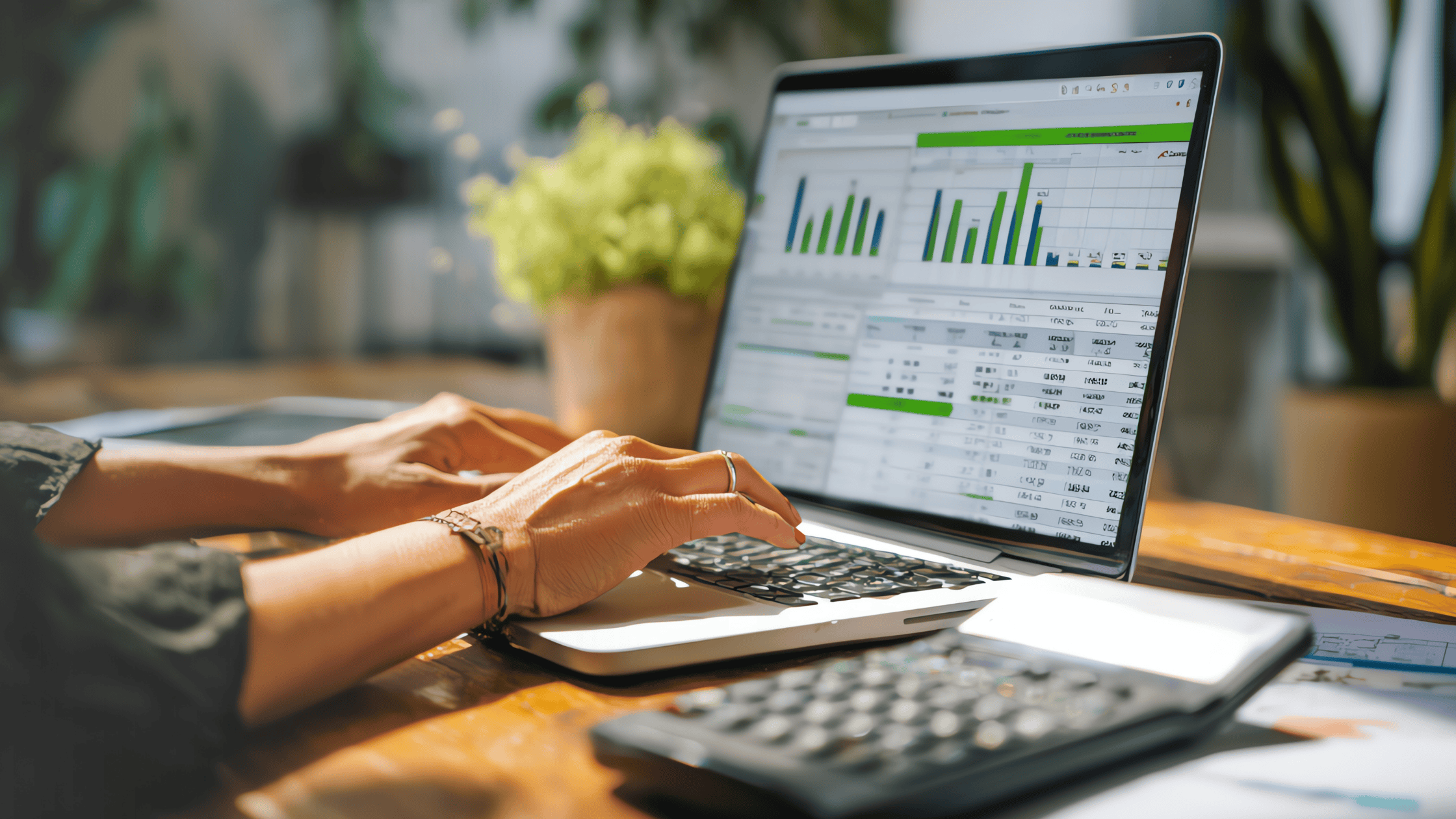 Hands typing on a laptop displaying spreadsheets and bar charts, with a calculator and documents on a wooden desk in a home or office setting.