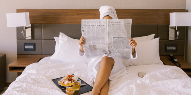 woman in hotel sitting on bed reading newspaper with room service beside her